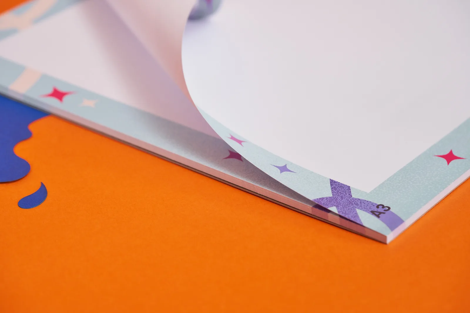 close up of an a3 printed desk pad with glue at the bottom edge with paper curled over on a bright orange background with blue paper ink spills