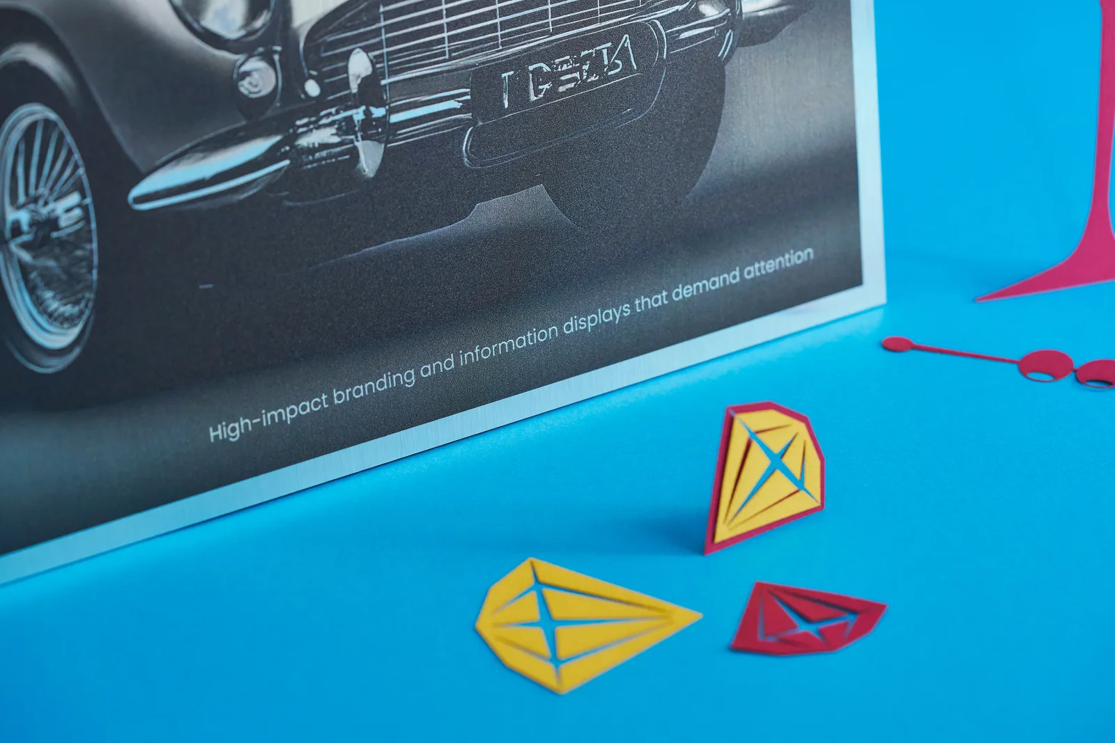 Close up of a brushed aluminium dibond sign with classic sports car on a bright blue background with yellow and purple paper diamonds