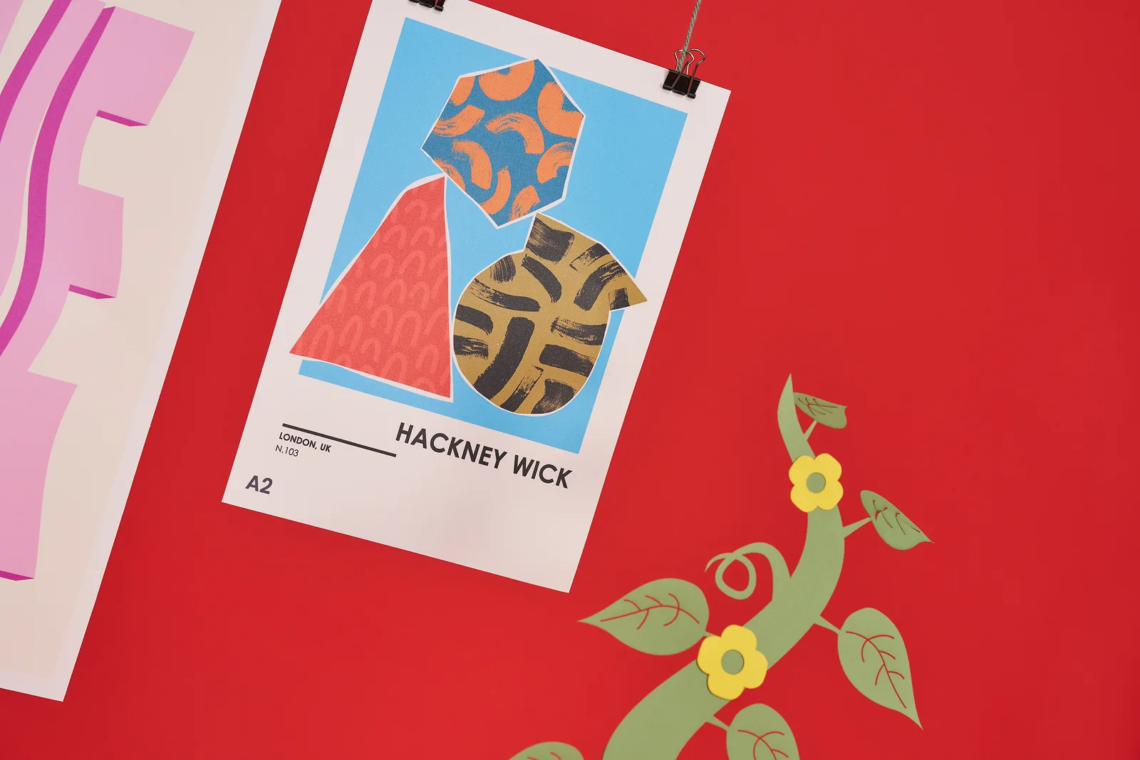 Close up of a giant A2 poster hanging from string with abstract art print with wording 'hackney wick' on a bright red background with green beanstalk and 2 yellow flowers