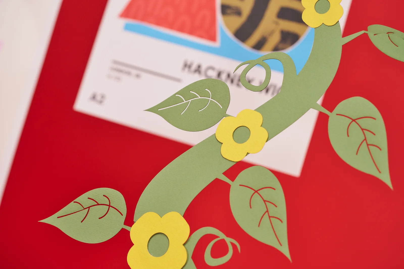 Close up of green paper beanstalk with 3 yellow flowers and behind is a A2 poster with abstract art on a bright red background
