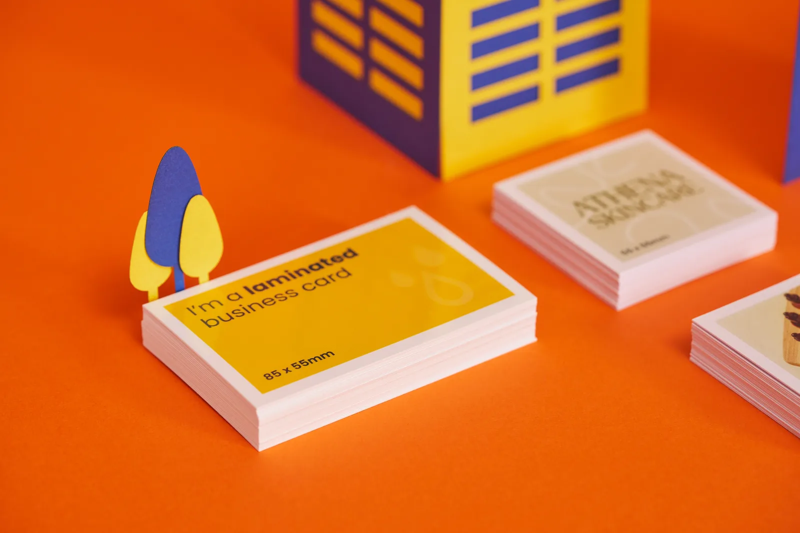 Close up of a pile of bright yellow 85mm x 55mm business cards with wording 'i'm a laminated business card'. 2 piles of 55mm x 55mm cream business cards with wording 'athena skincare' on a b