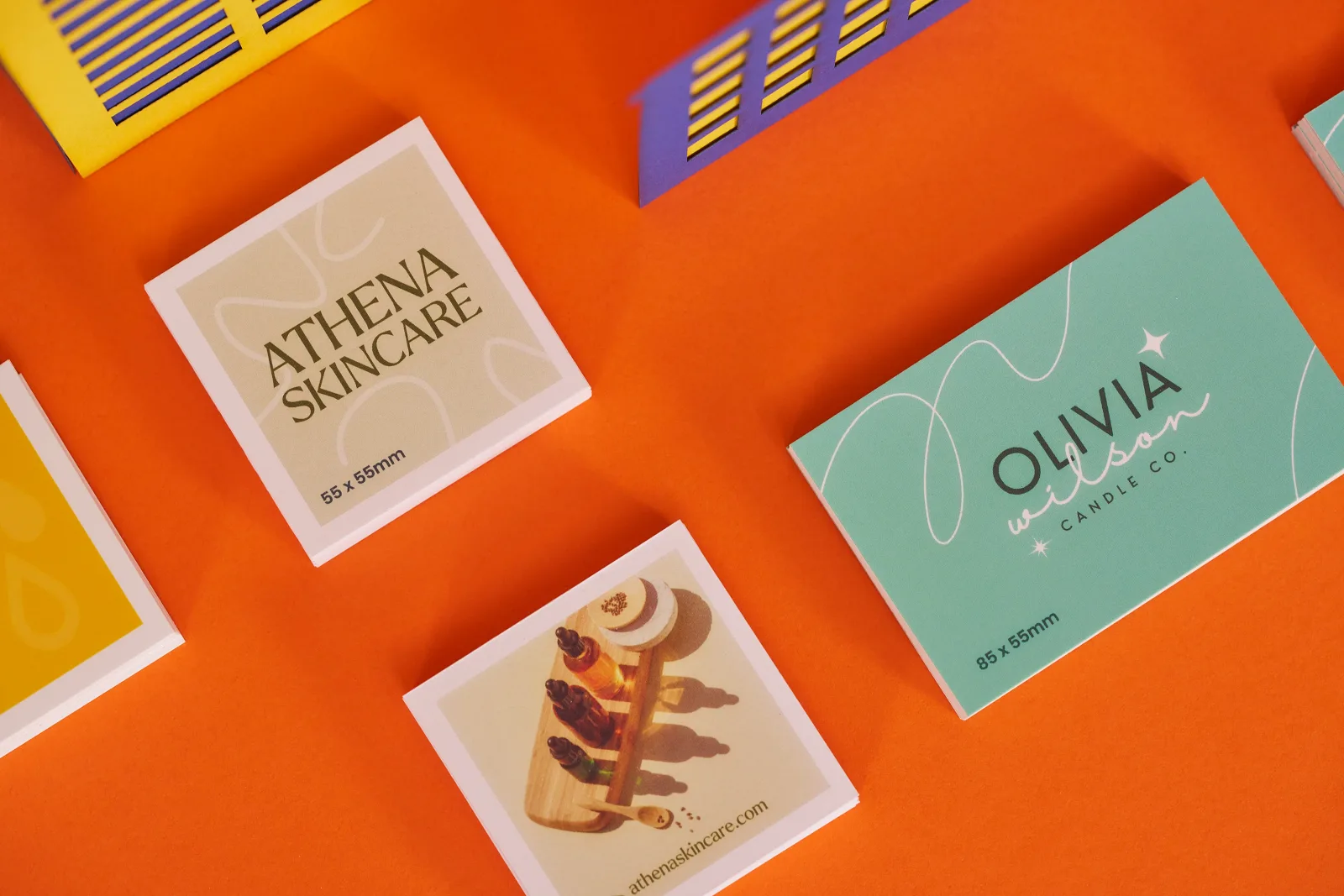 Close up of 2 piles of 55mm x 55mm cream business cards with wording ' athena skincare' and 1 pile of mint green 85mm x 55mm business cards with working 'olivia wilson' on a bright orange ba