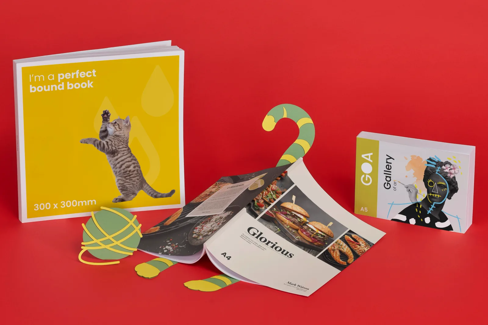 3 printed perfect bound books 300 x 300mm, A4 and A5. A4 book has a fun paper cat hiding underneath with paws and tail sticking out chasing a paper ball of wool on bright red background