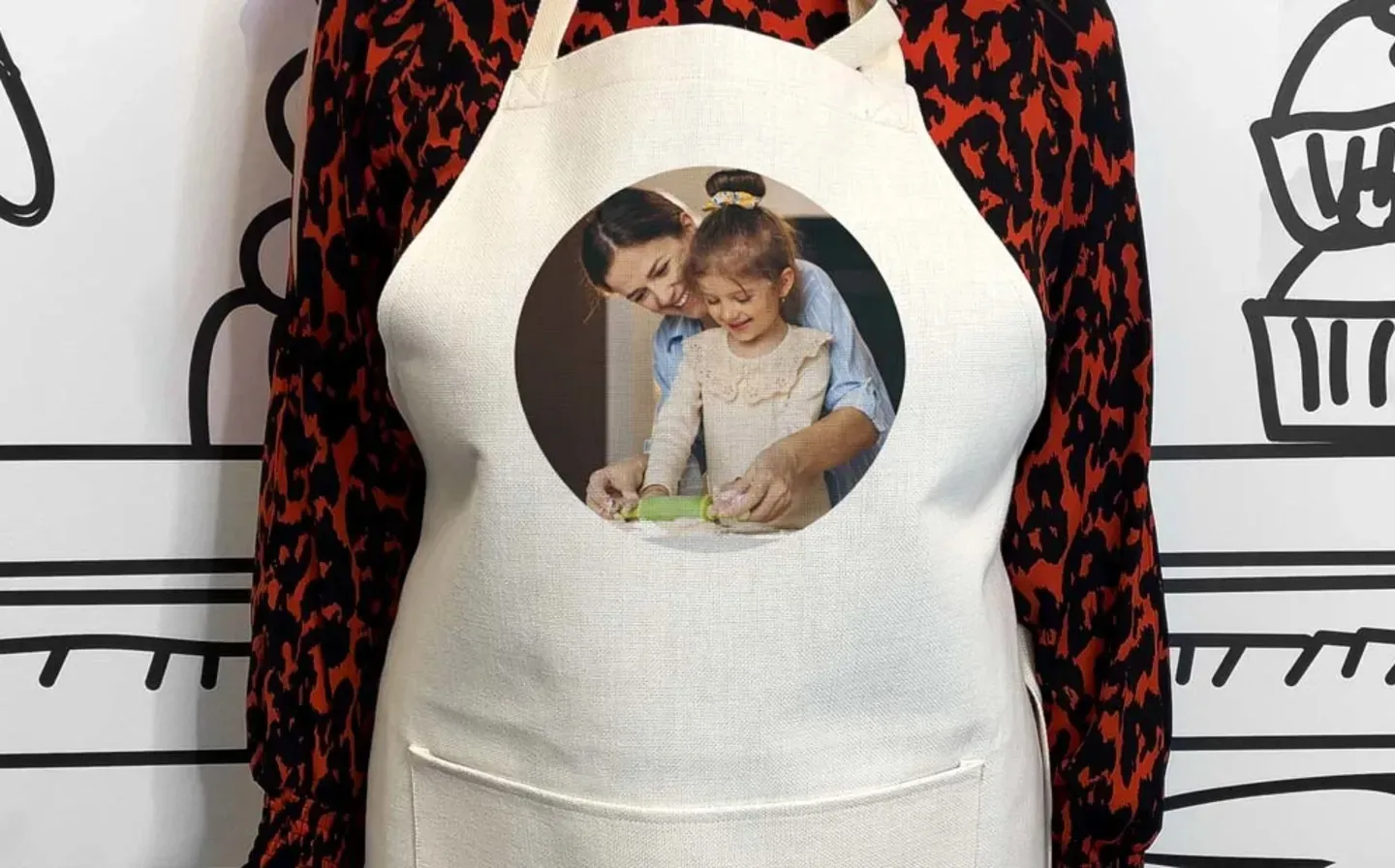  A woman stands in a kitchen, wearing an apron featuring a photograph of a woman and child