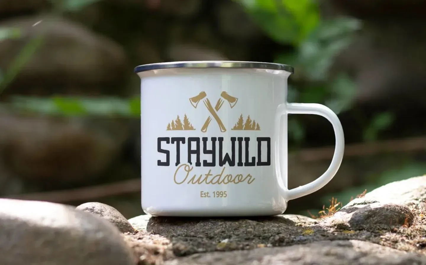 A stylish enamel mug featuring a "Stay Wild Outdoors" logo, displayed on an outdoors scene