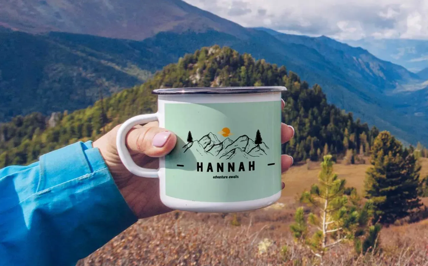 A woman holding an enamel mug inscribed with "Hannah", set against a stunning backdrop of mountains