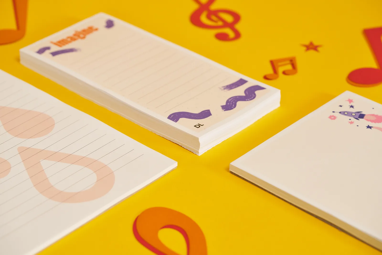 close up of 3 printed notepads with various number of pages, dl portrait with text 'imagine', a5 portrait with rocket ship illustration on a yellow background with orange and red paper music