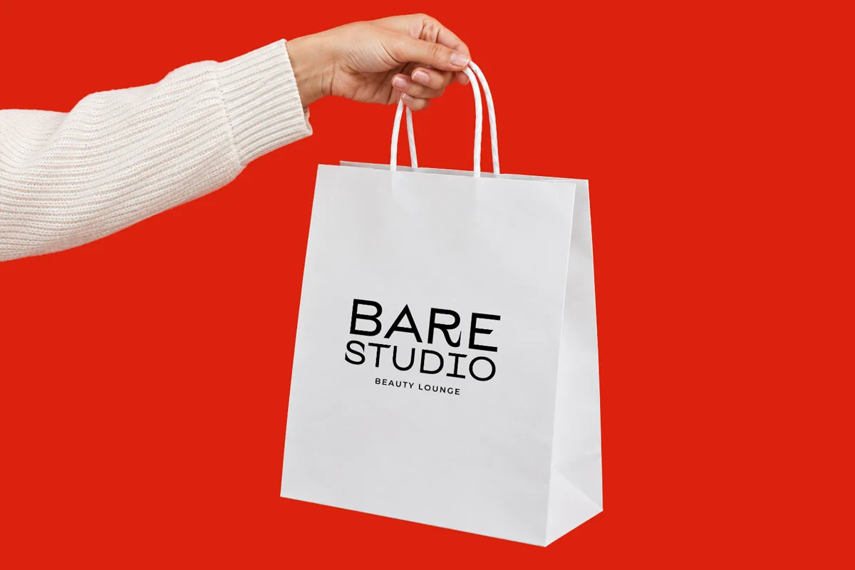A hand holding a white shopping bag branded with the Bare Studio Beauty Lounge logo.
