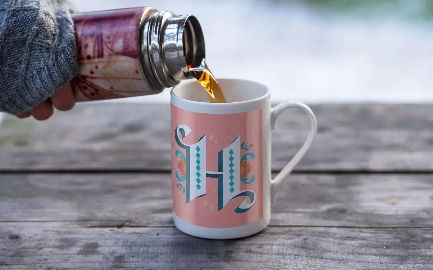 A person pouring coffee into a porcelain mug from a flask, adorned with a stylish "H" design