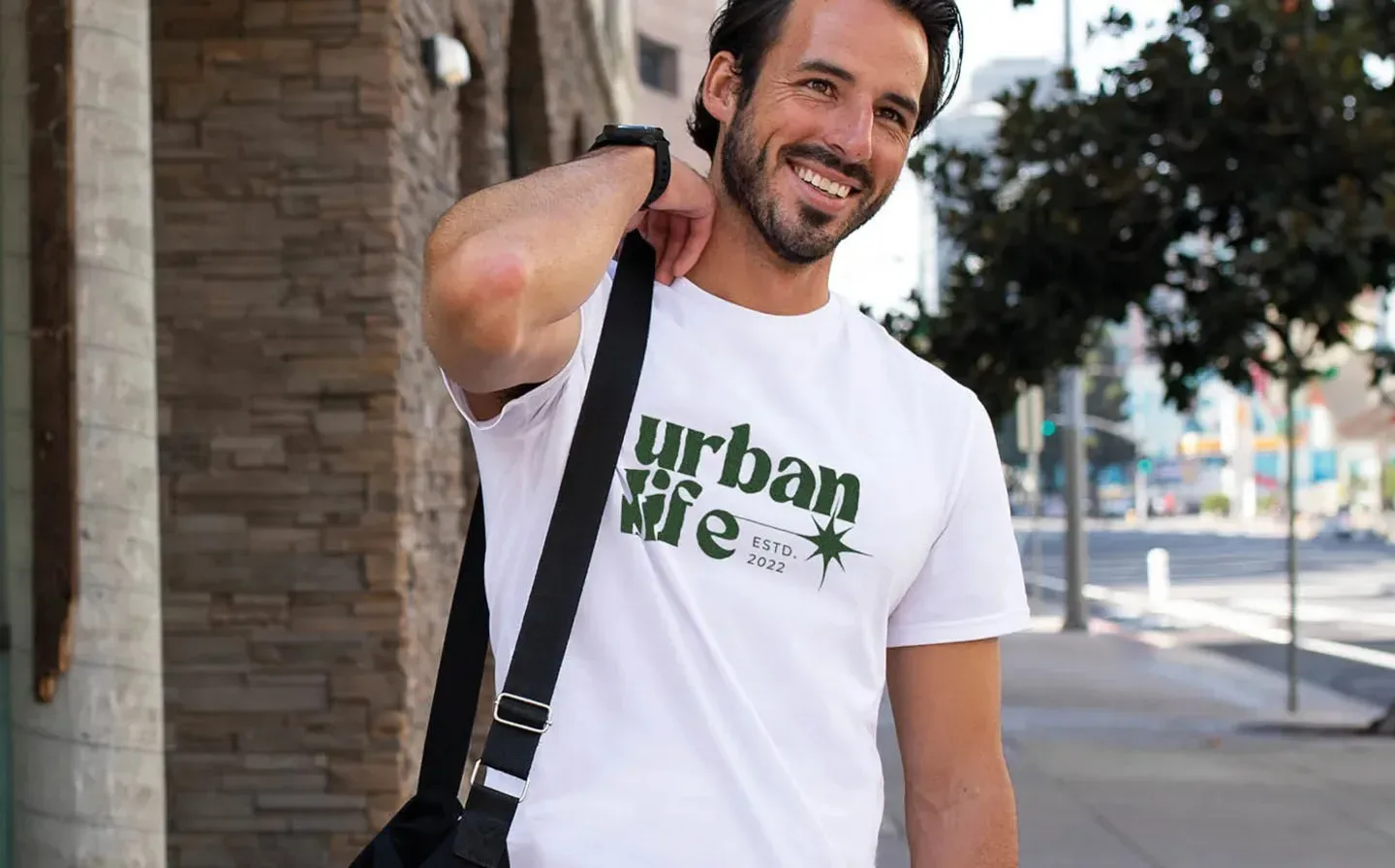 A man in a white t-shirt displaying the phrase "urban life," standing against a city backdrop