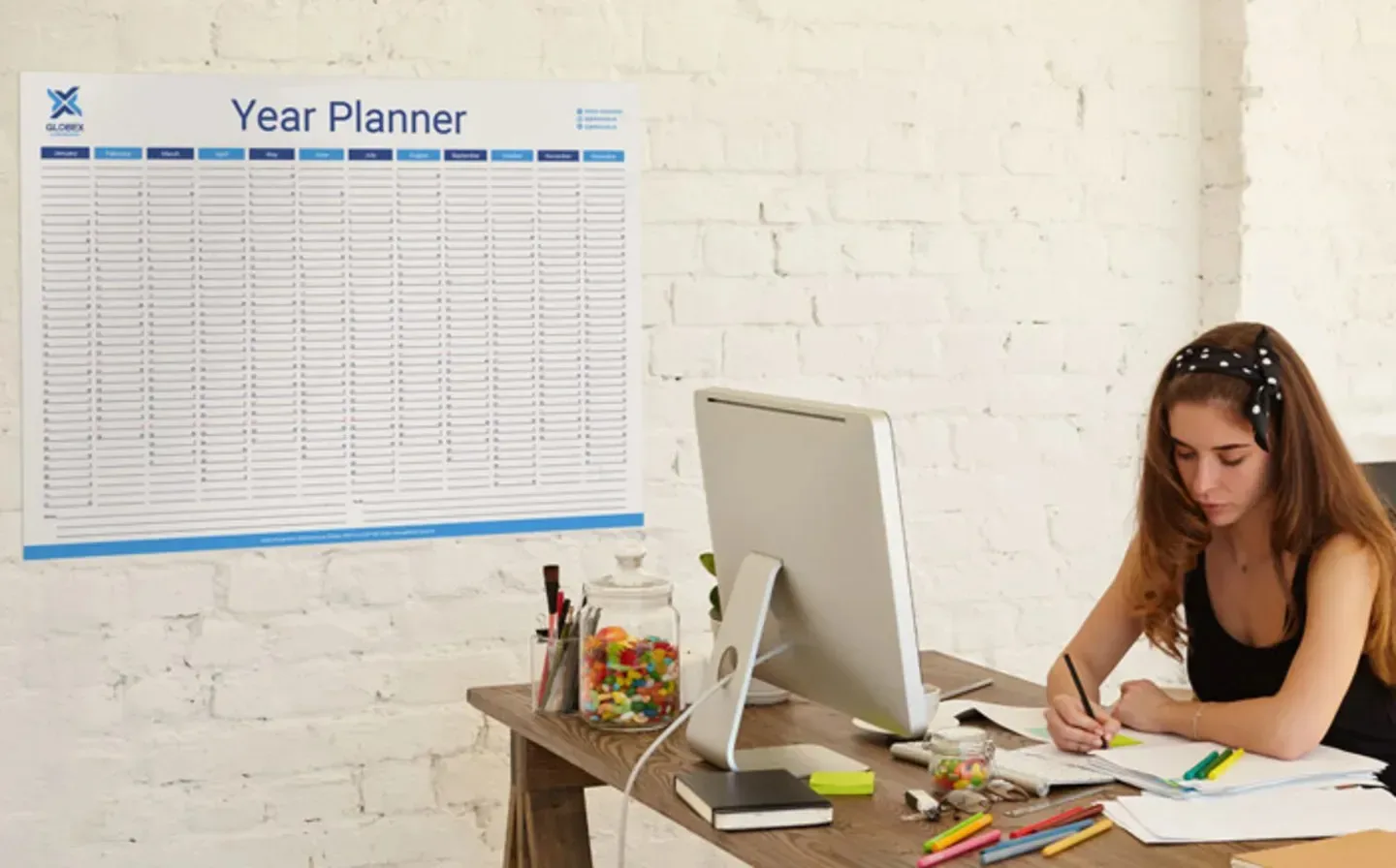 A woman seated at a desk with a wall planner prominently displayed behind her
