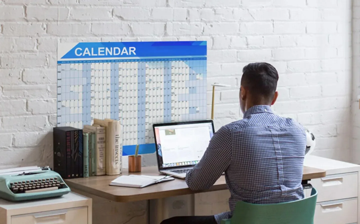 A man seated at a desk, focused on work, with a wall planner visible behind him