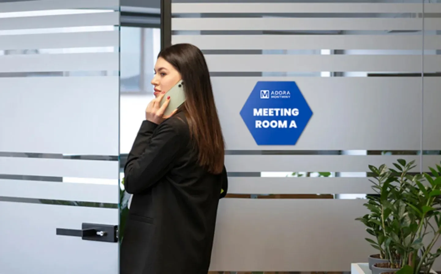 A woman on her cell phone, standing in front of a glass door with a photo tile, showing "Meeting Room A"