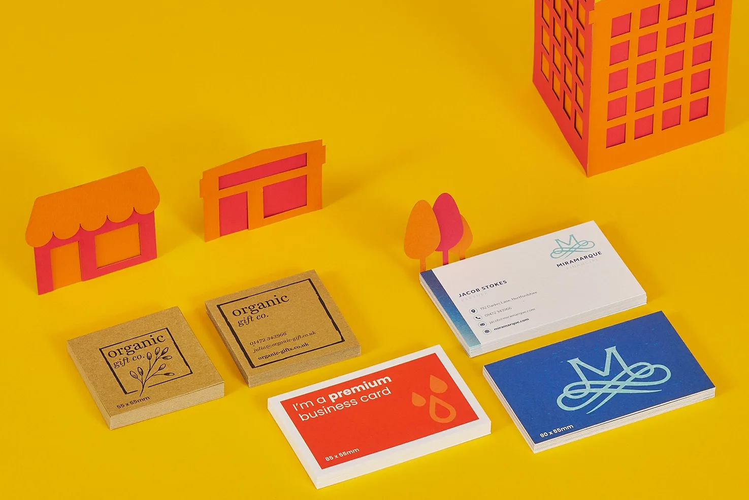 5 stacks of premium on business cards with copy 'i'm a premium business card 85 x 55mm' and 'organic gift.co 55 x 55mm on a bright yellow background with orange and red paper buildings and t