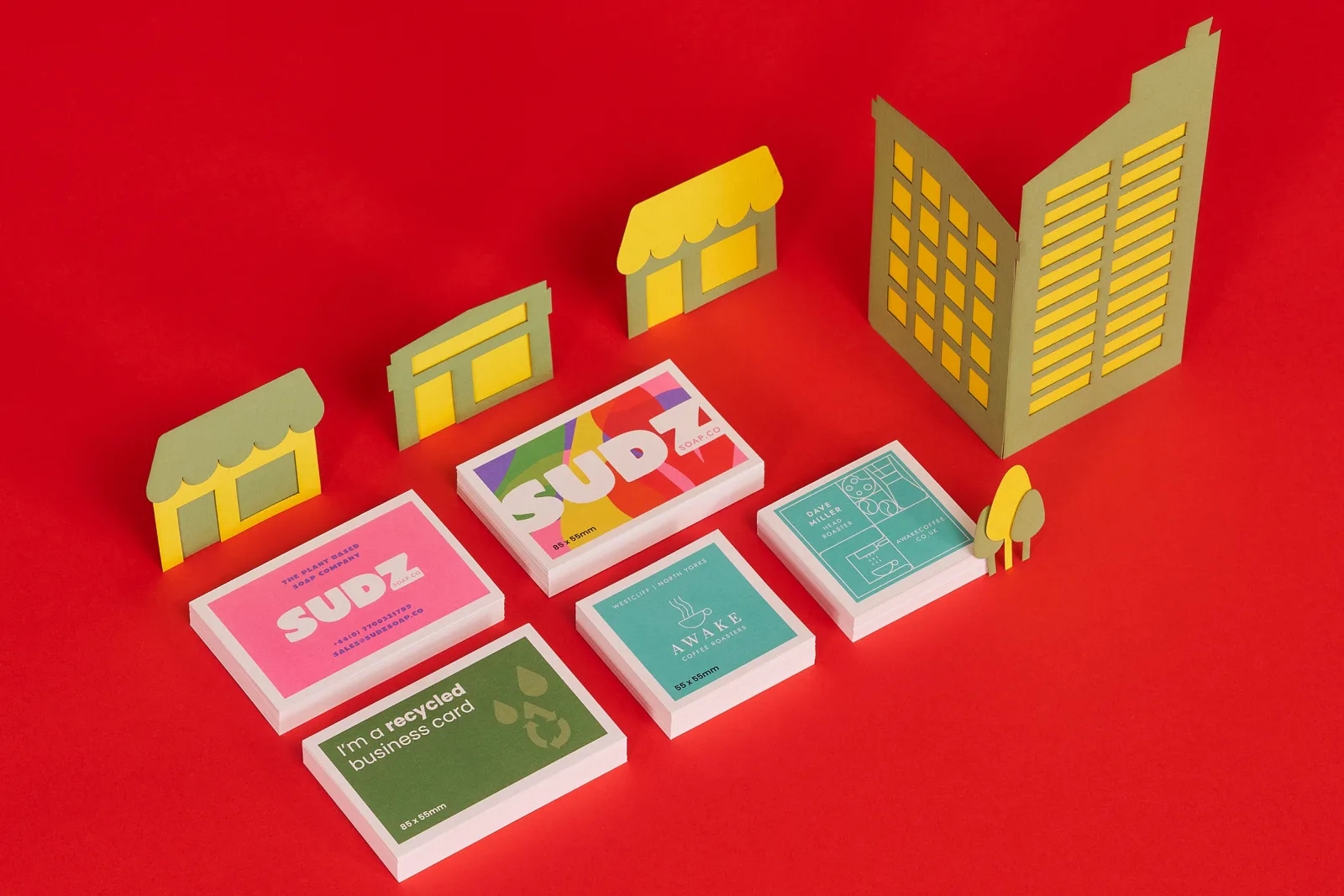 stack of 5 recycled business cards. Pink with text 'sudz', rainbow with 'sudz', green with 'i'm a recycled business card' on a bright red background with green and yellow paper buildings and