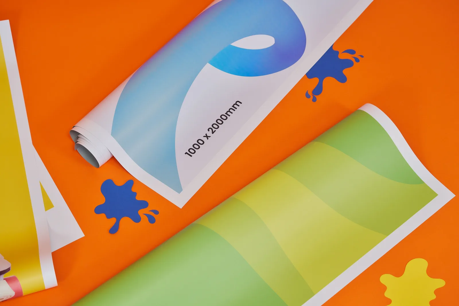 replacement roller banner graphics rolled up one green and one blue with text '1000 x 2000mm' on a bright orange background with yellow and blue paper ink spills
