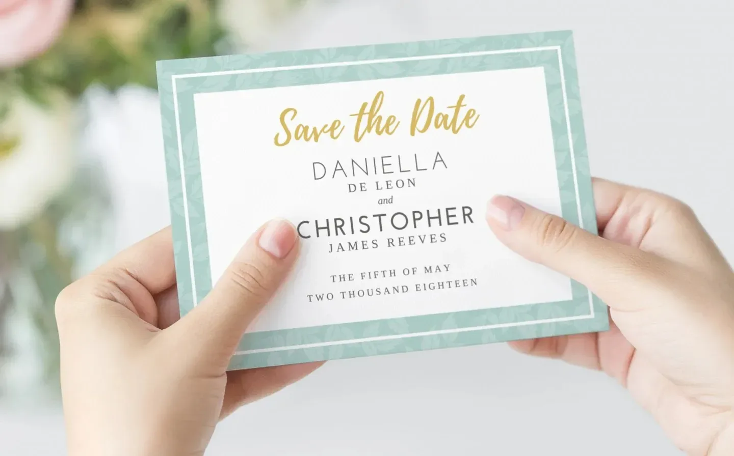 A hand holds a wedding save the date card in front of a beautiful flower arrangement