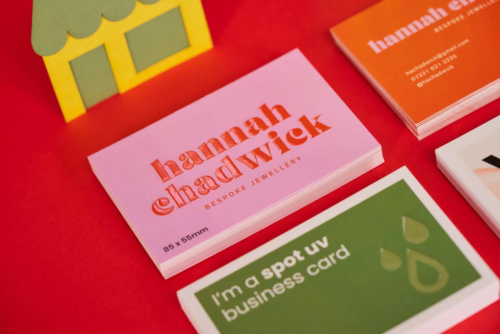 4 stacks of business cards with text 'hannah chadwick' in red writing with spot UV overlay. Green business card with spot uv and text 'i'm a spot uv business card' on a bright red background